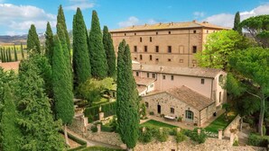 A historic building with a courtyard surrounded by tall cypress trees, stone walls, and a tiled roof.