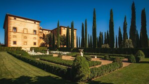 A large, historic building with a symmetrical facade, surrounded by well-manicured gardens and tall cypress trees.
