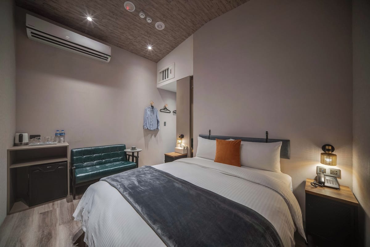 Een moderne hotelkamer met een bed, nachtkastje met lamp, bureau met stoel, garderobe en een airconditioning.
