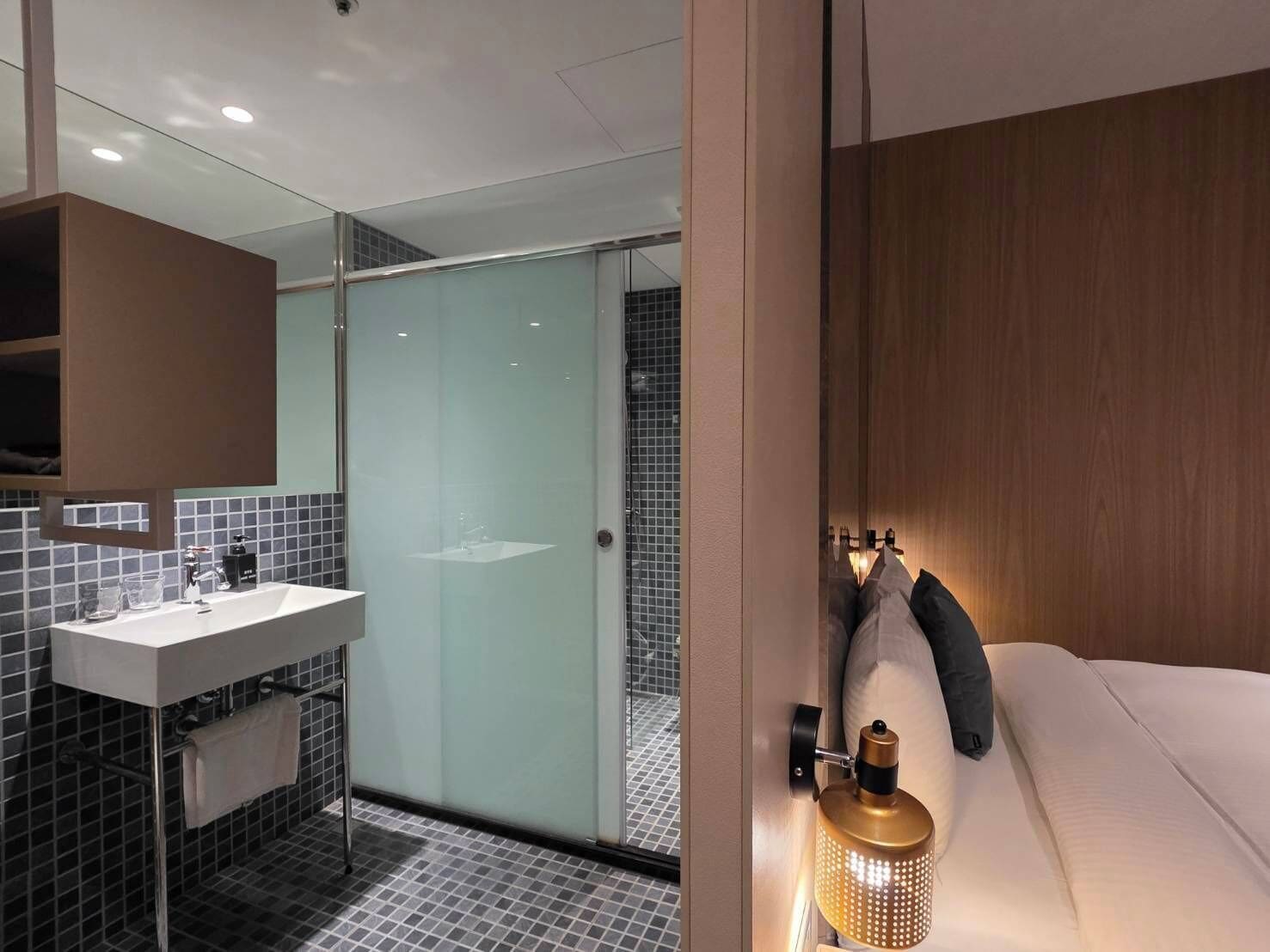 A modern bathroom with a glass shower enclosure, a white sink, and a bedside lamp.