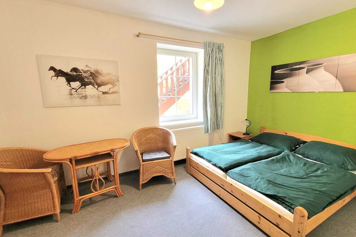 A room with a bed, two wicker chairs, a small table, a window with curtains, and two wall-mounted pictures.