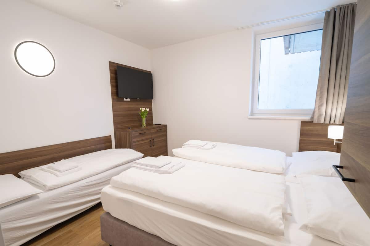 Een hotelkamer met drie enkele bedden, een houten hoofdbord, een flatscreen televisie aan de muur, een raam met gordijnen en een nachtkastje met een bloevas.