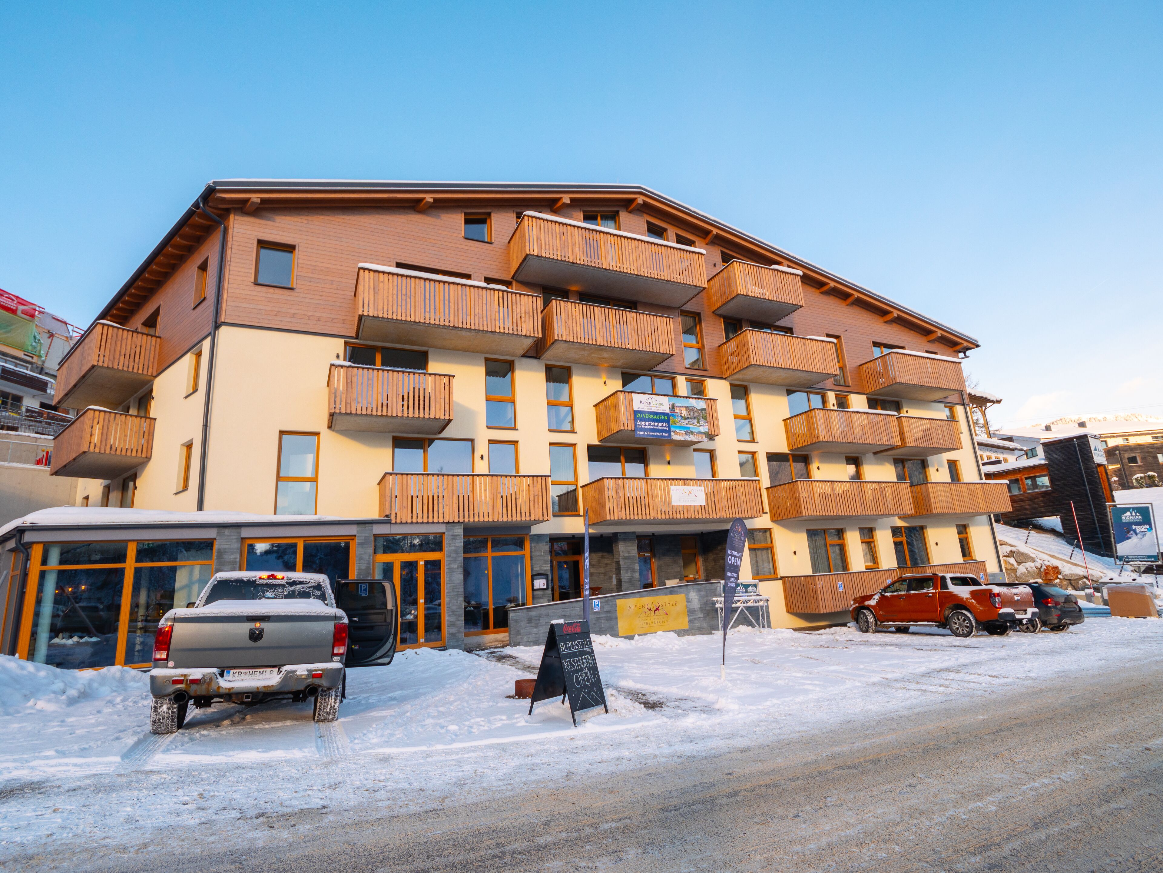 Een meerlaags hotel met houten balkons, een restaurant en een parkeerplaats met sneeuw.