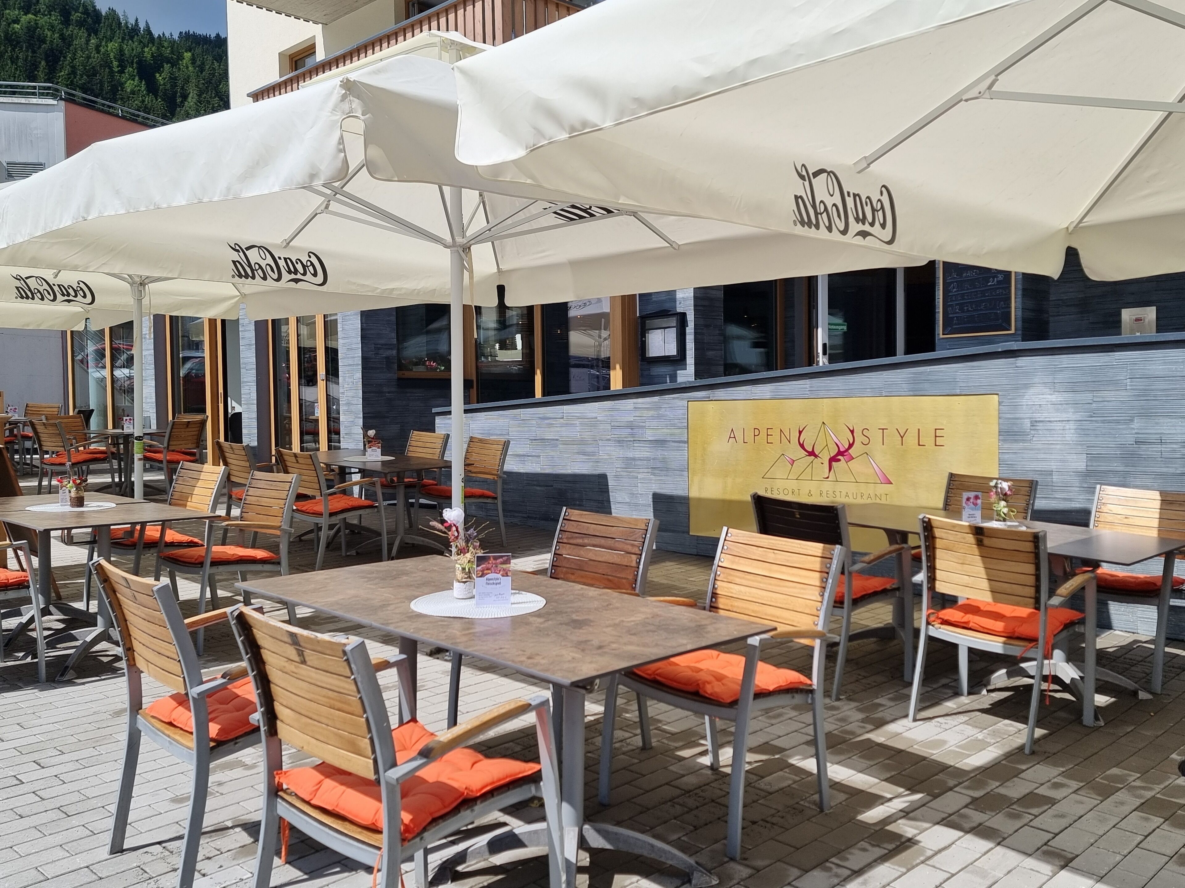 An outdoor dining area with tables and chairs, umbrellas, and a sign for AlpenStyle Resort & Restaurant.