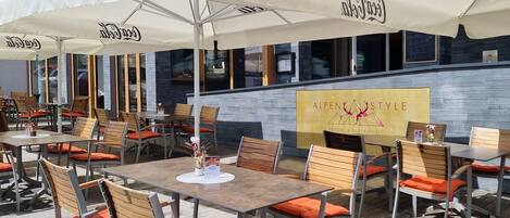 An outdoor dining area with tables and chairs, umbrellas, and a sign for AlpenStyle Resort & Restaurant.