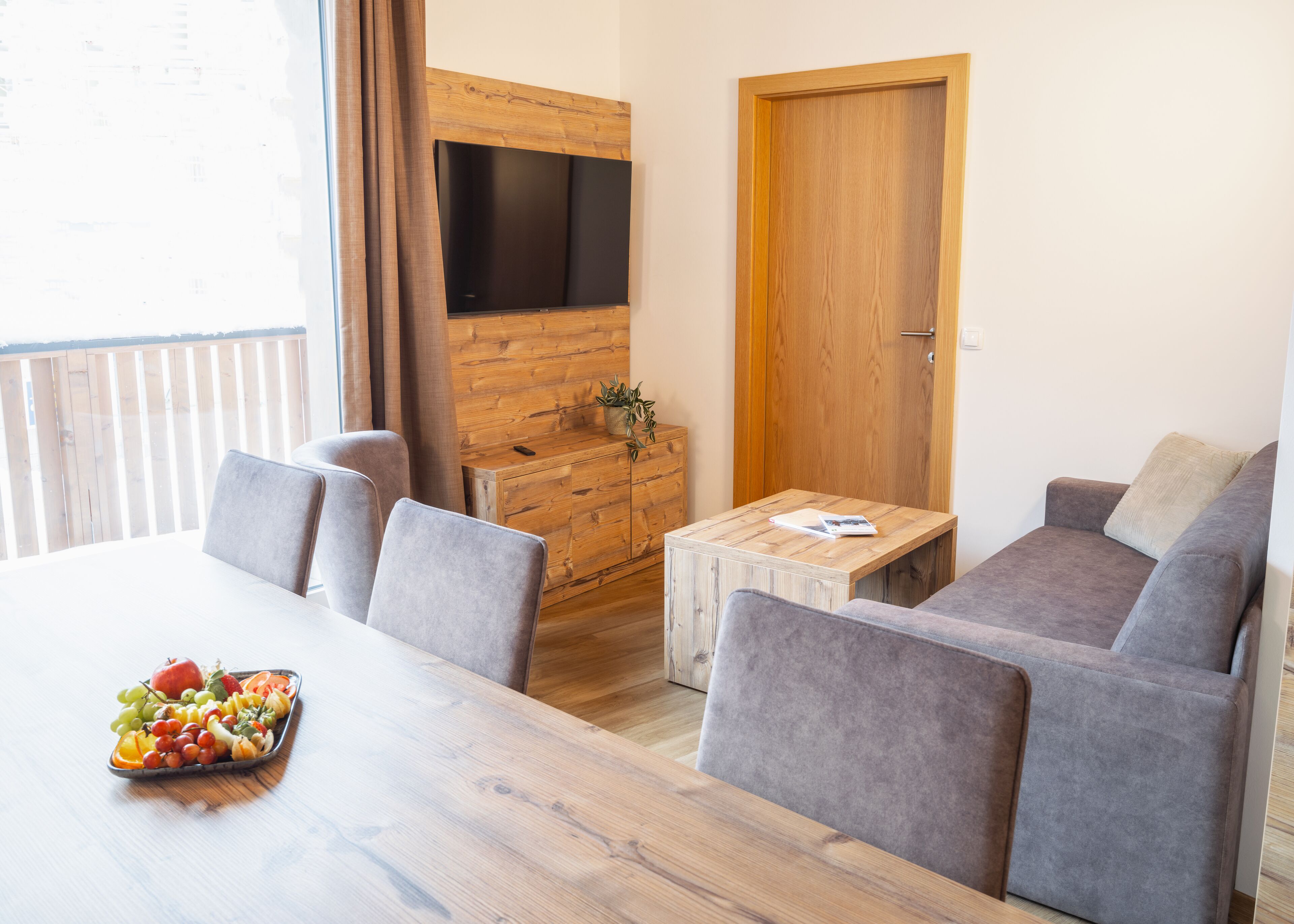 A modern living room with a wooden TV stand, grey sofas, a wooden coffee table, and a fruit platter on the dining table.