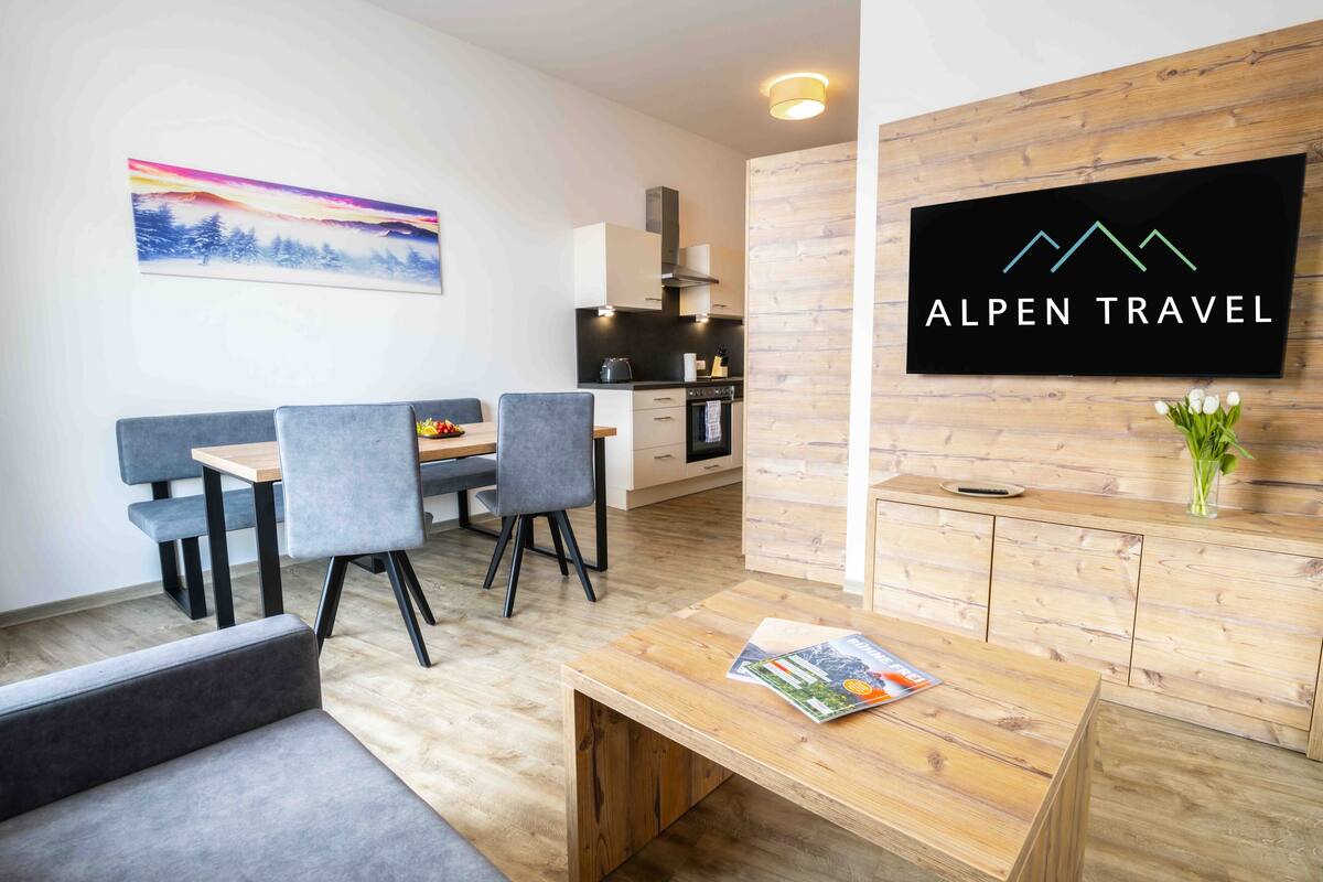Een moderne hotellobby met een houten receptiebalie, een zithoek met grijze fauteuils, een eettafel met een fruitschaal, en een keuken op de achtergrond.