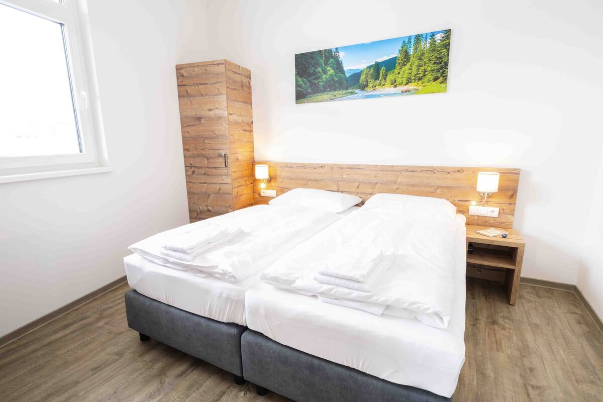 Een slaapkamer met een bed, een houten hoofdbord, twee nachtkastjes, een wandlamp en een lijst met een landschapsfoto.