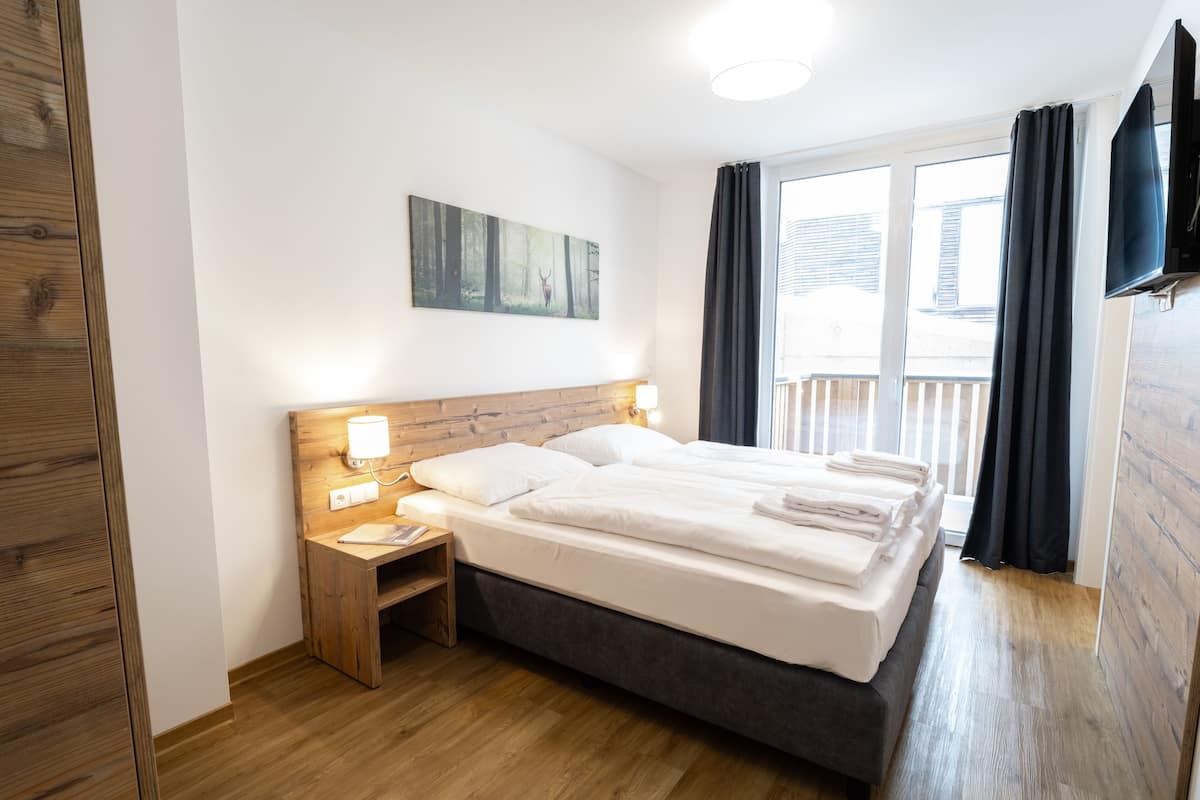 Een hotelkamer met een houten vloer, een bed met witte lakens, een nachtkastje met een lamp, een schilderij aan de muur en een raam met gordijnen.