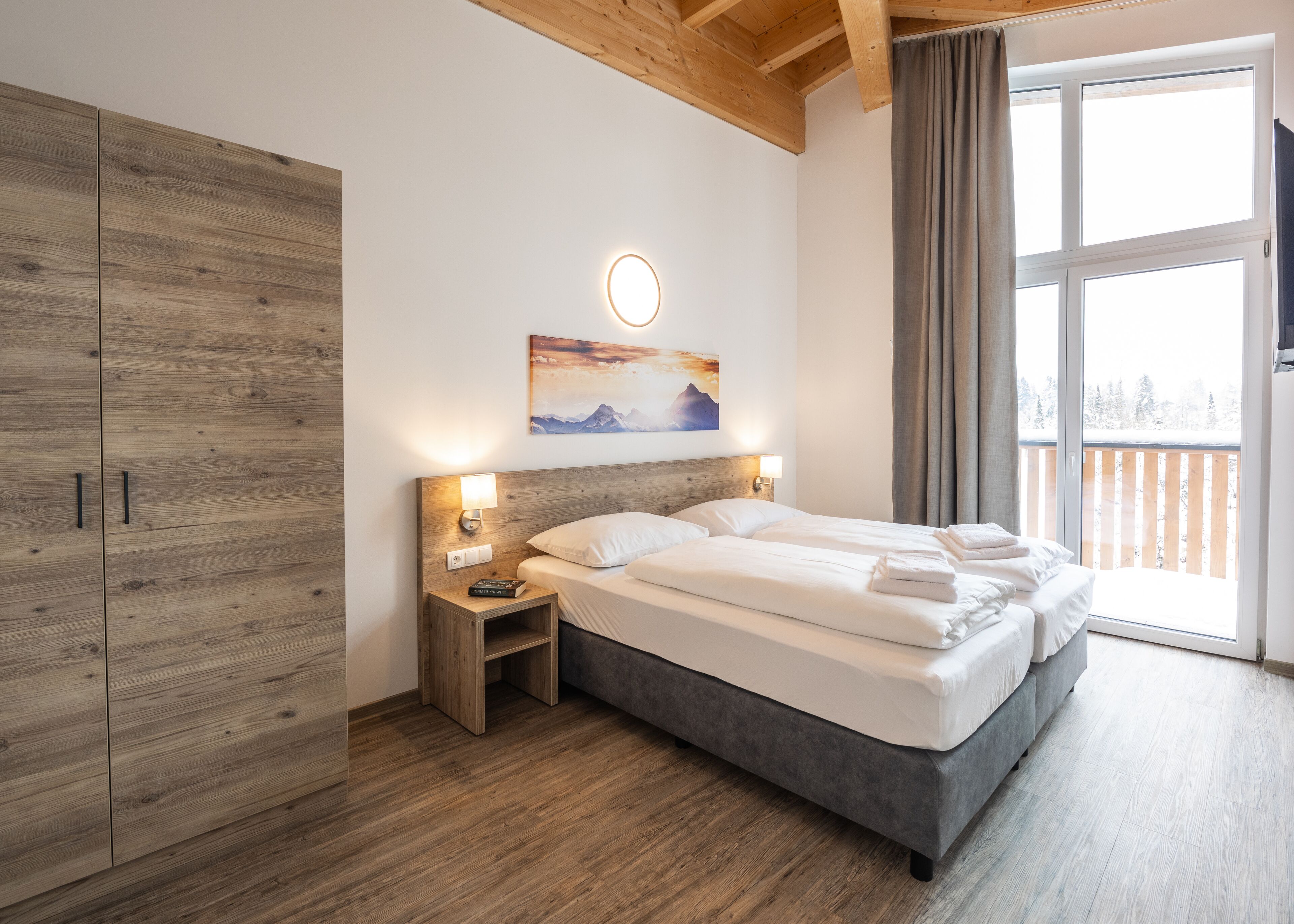 Une chambre à coucher avec un lit, des tables de chevet, une armoire en bois, un tableau et une vue sur un paysage enneigé par une grande fenêtre.