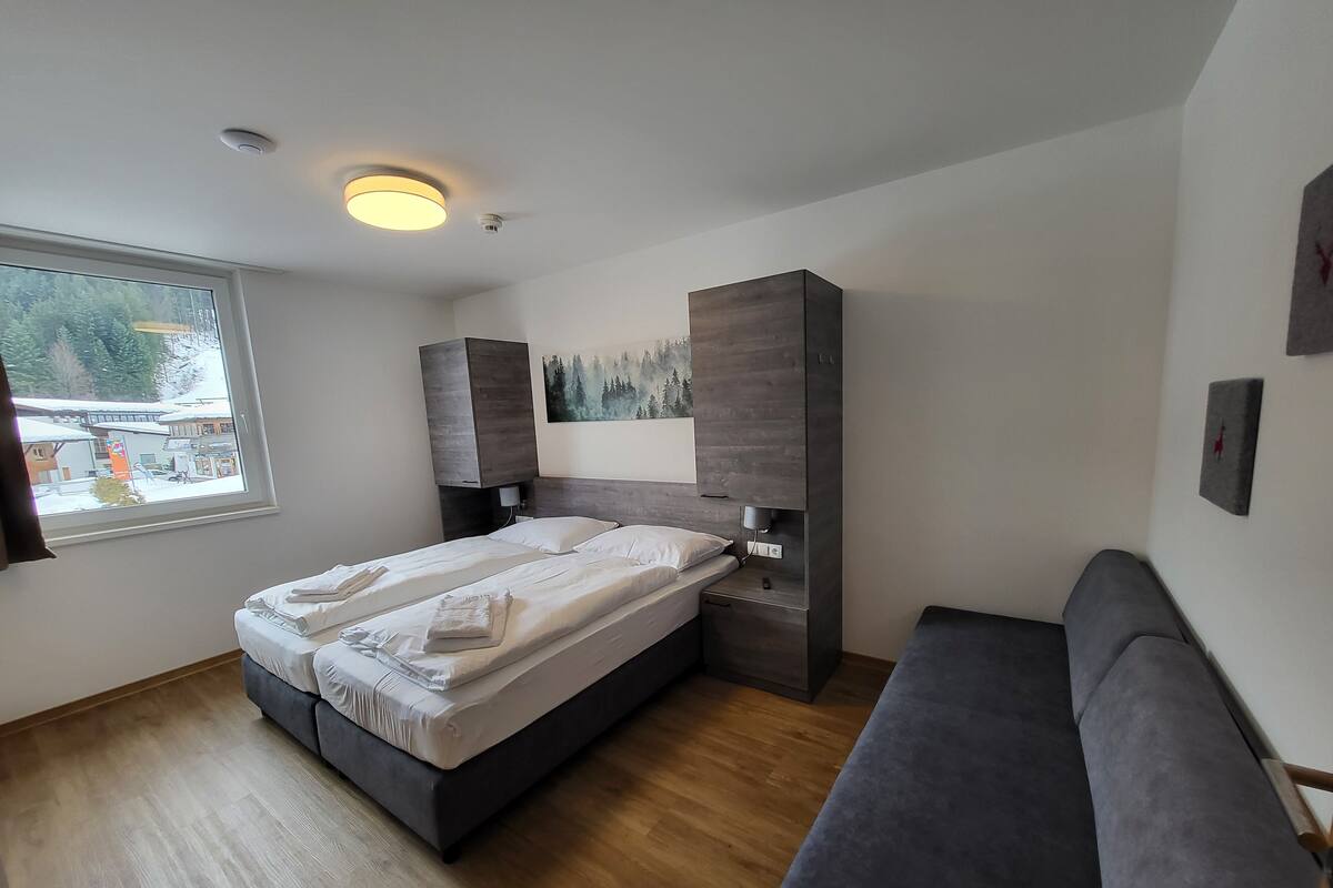 Een moderne slaapkamer met een bed, een inbouwkast, een grijze bank en een raam met uitzicht op gebouwen en bomen.