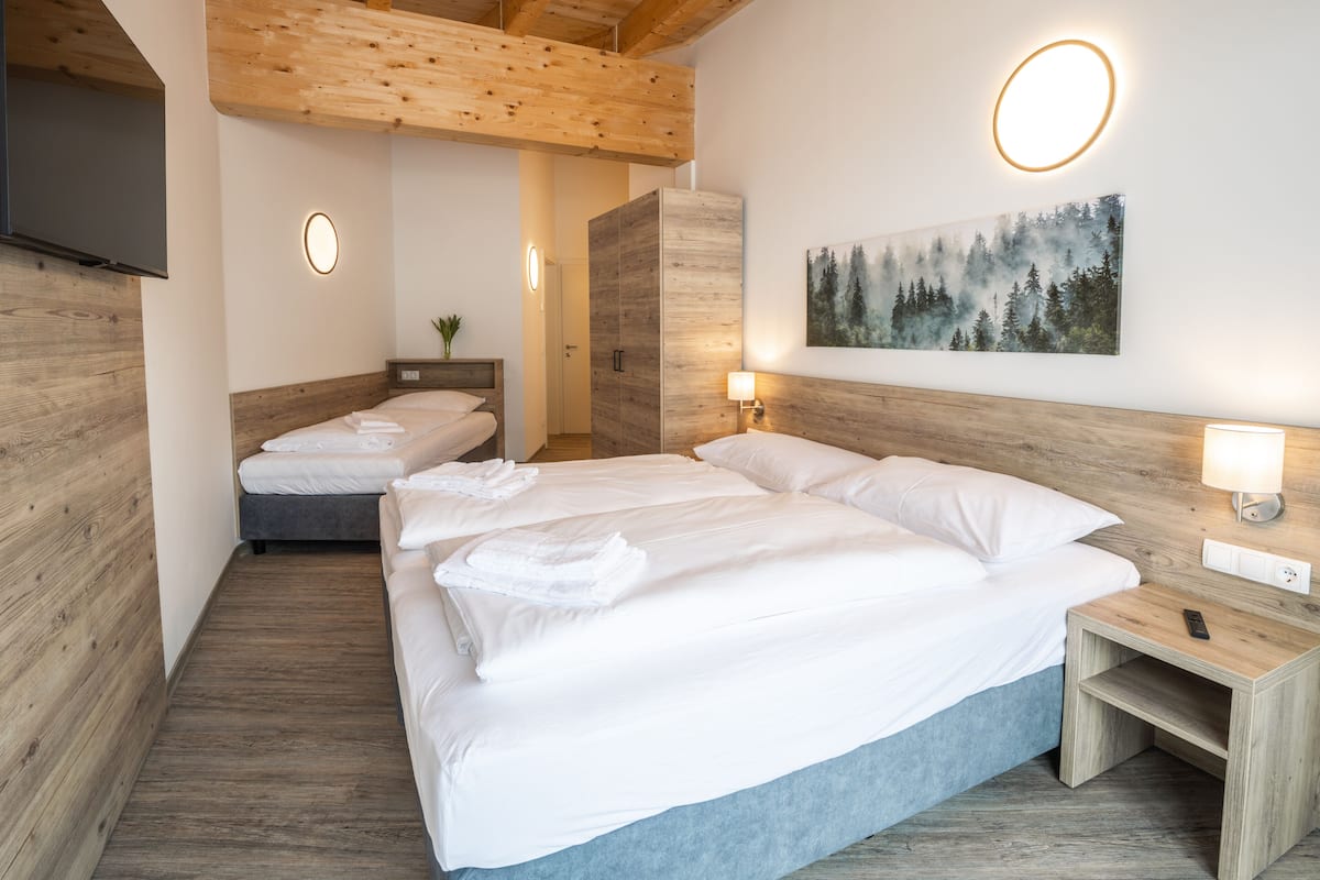 Een hotelkamer met een houten plafond, een schilderij van een bos, twee bedden met witte lakens en een nachtkastje met een lamp.