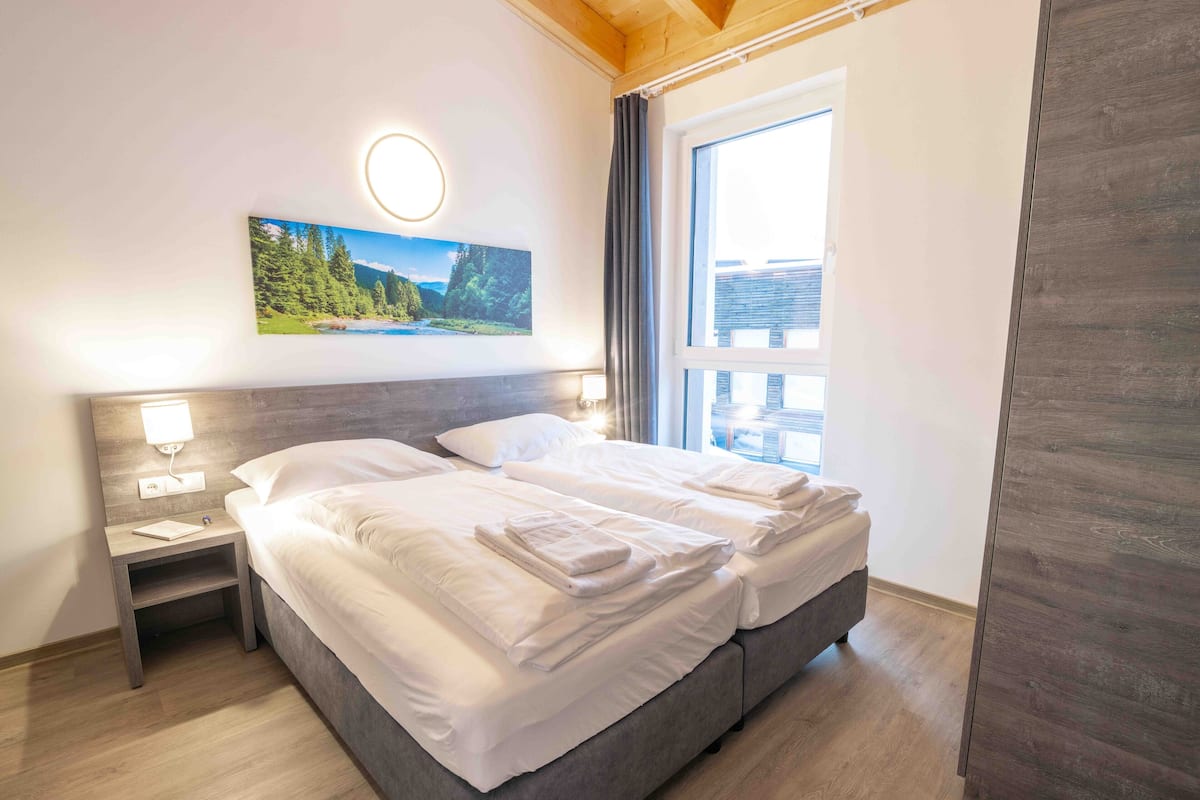 Een keurig opgemaakt bed met witte lakens en handdoeken, een nachtkastje met een lamp, een ingelijst beeld aan de muur, en een groot raam met gordijnen.