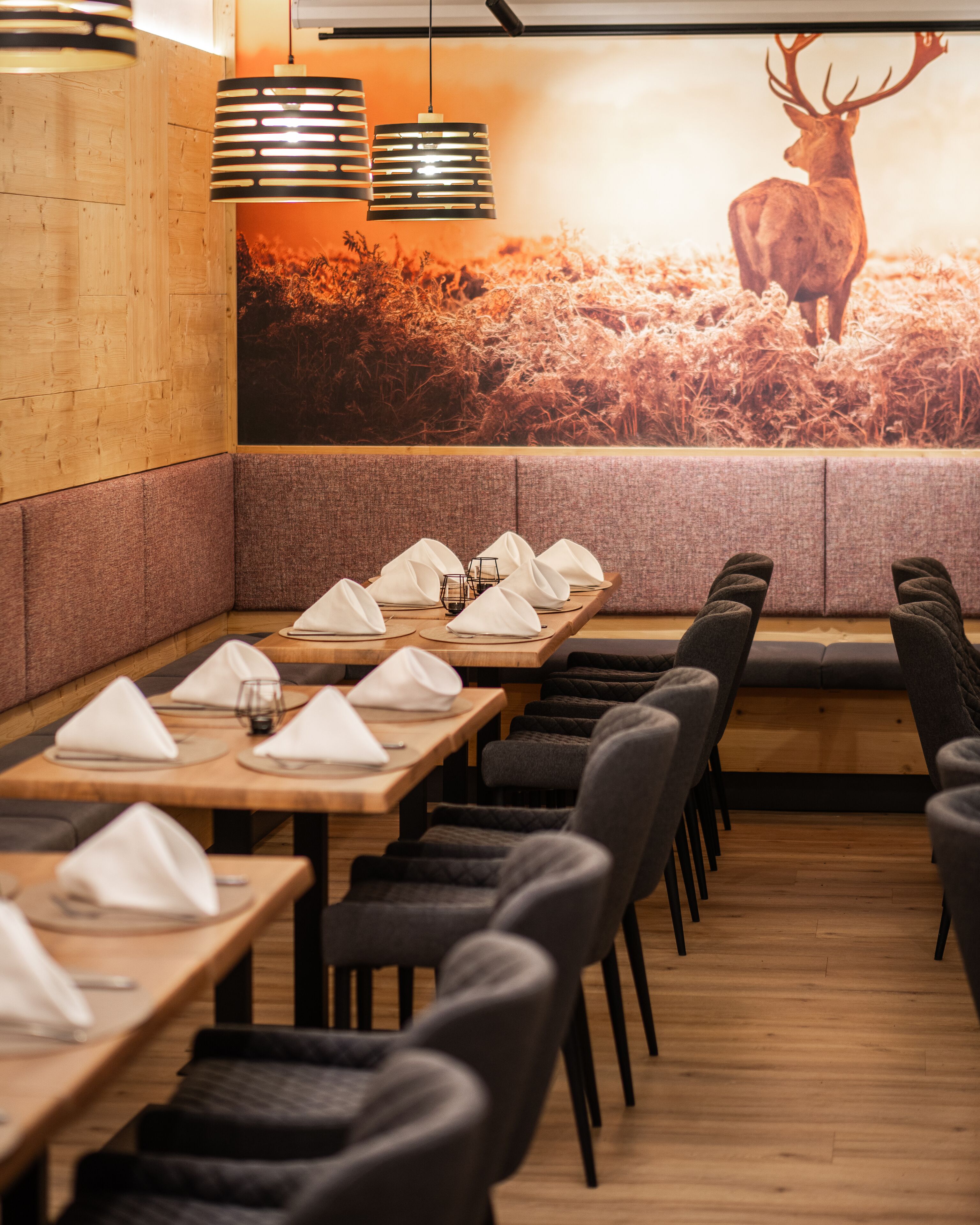 A restaurant with wooden tables and black chairs, a mural of a deer, and pendant lighting.