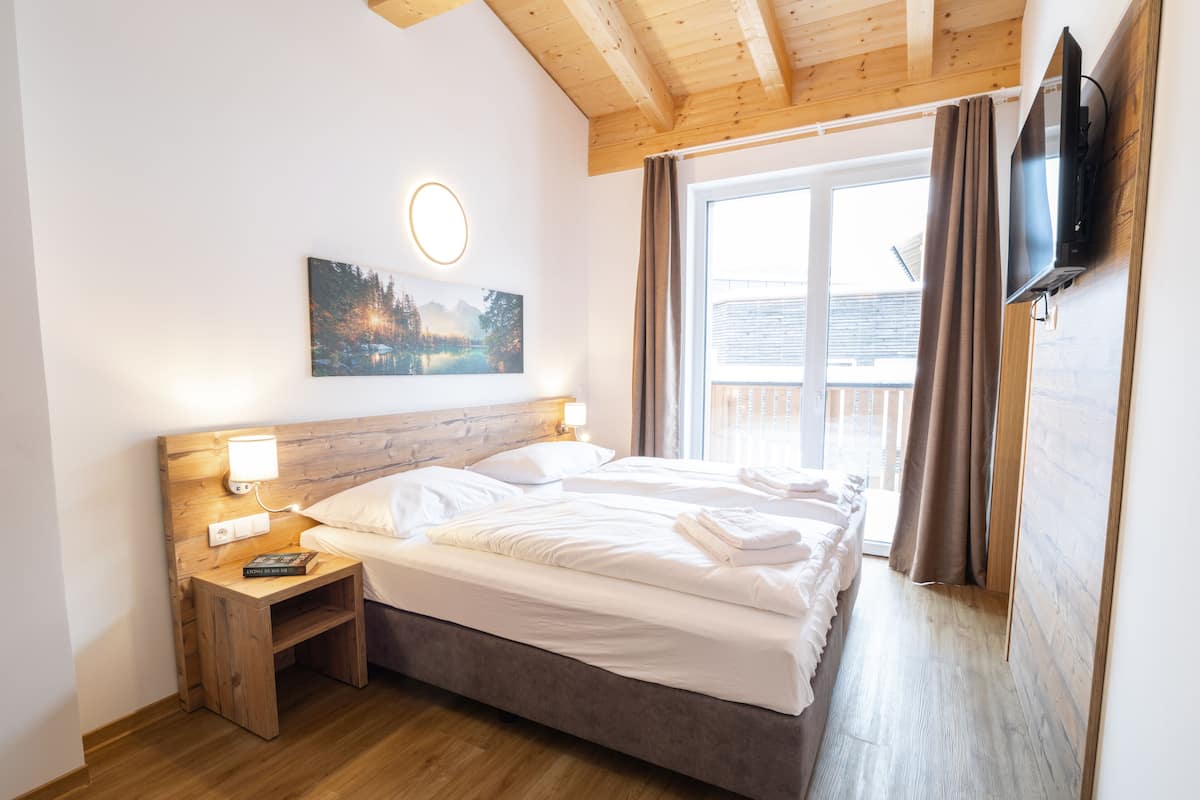 Een slaapkamer met een houten hoofdbord, een bed met witte lakens, een nachtkastje met een lamp, een schilderij aan de muur en een aan de muur bevestigde televisie.
