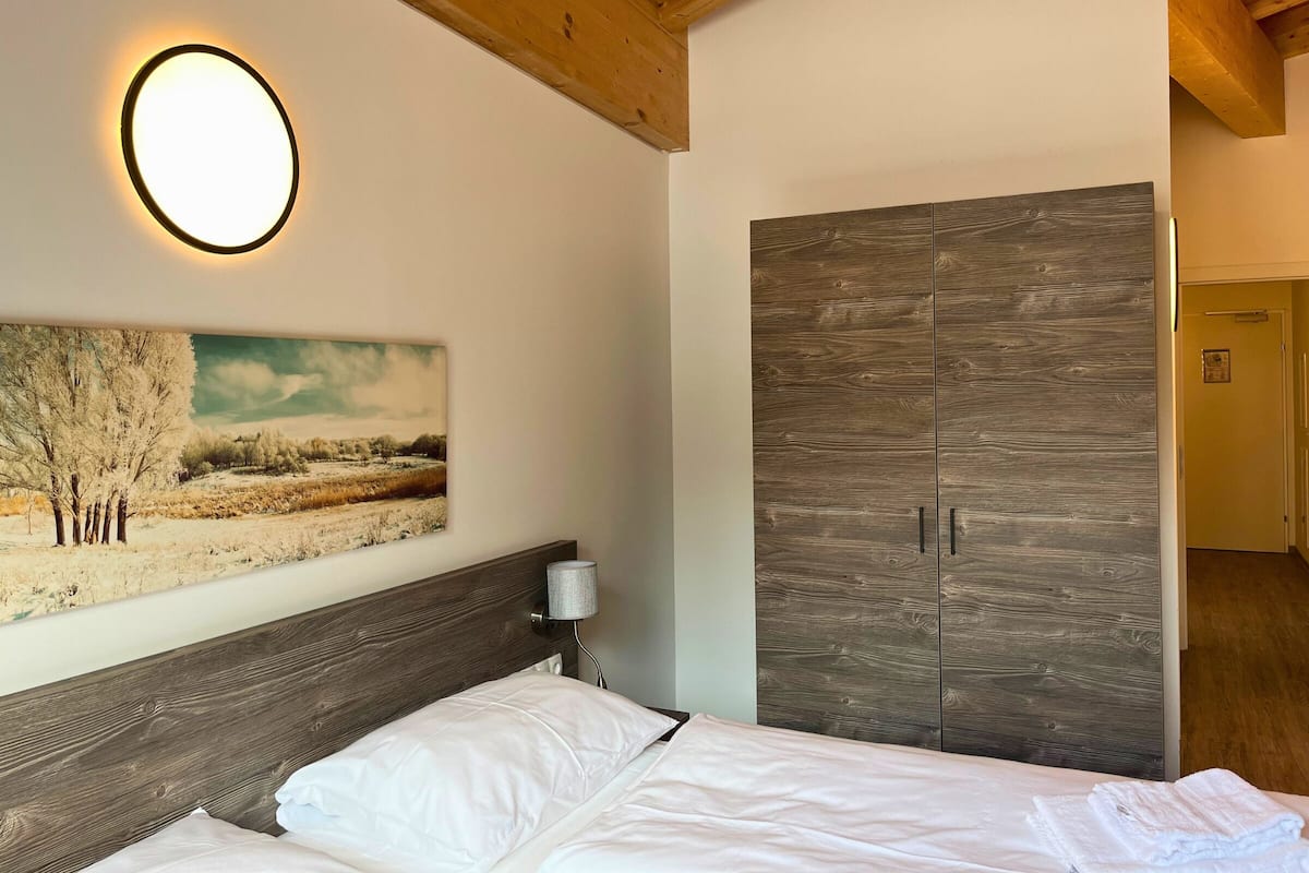 Een slaapkamer met een bed, een houten garderobe, een schilderij aan de muur en een nachtkastje met een lamp.
