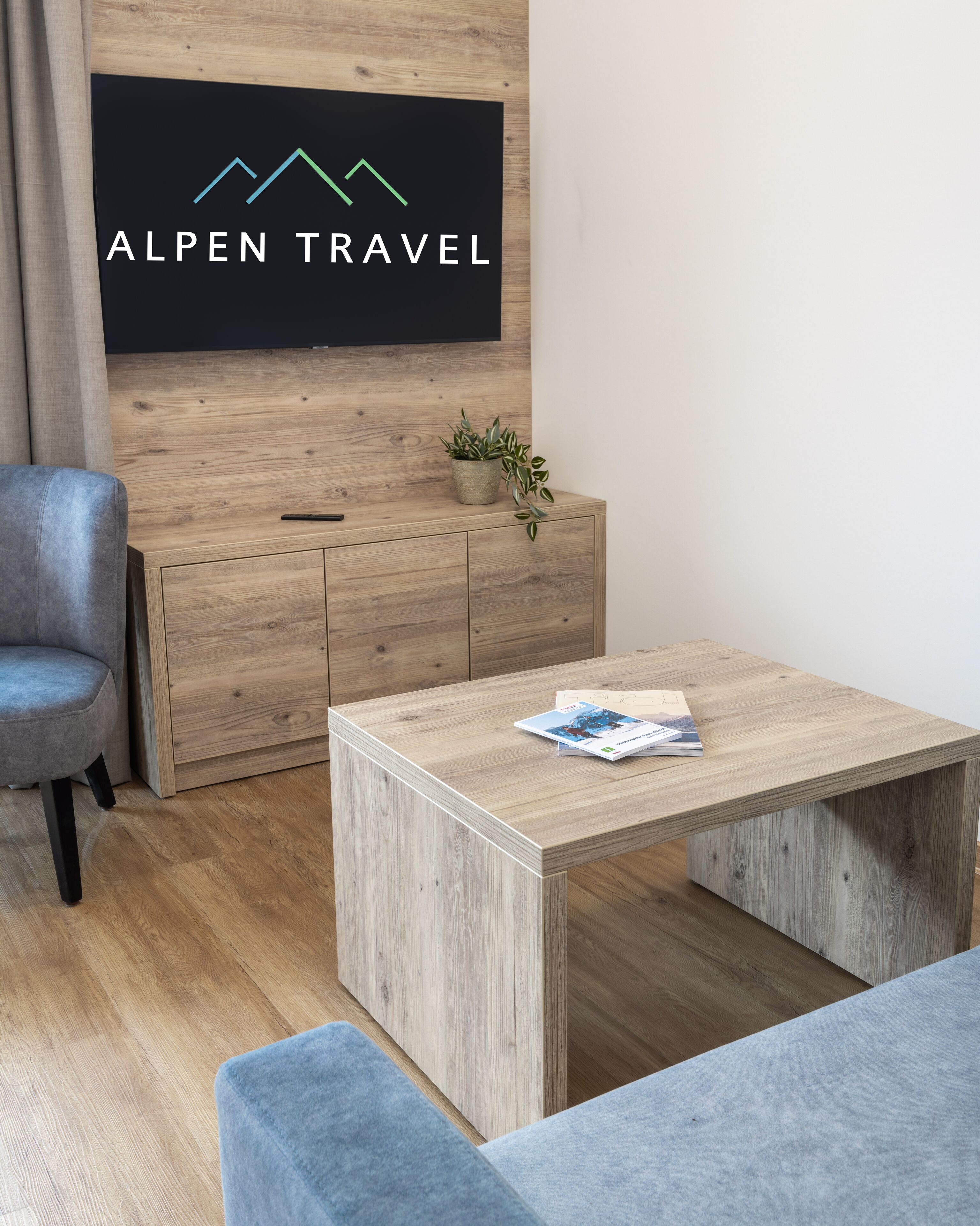 A modern room with a wooden cabinet, a grey sofa, a wooden coffee table with a magazine, and a wall-mounted TV displaying 'ALPEN TRAVEL'.