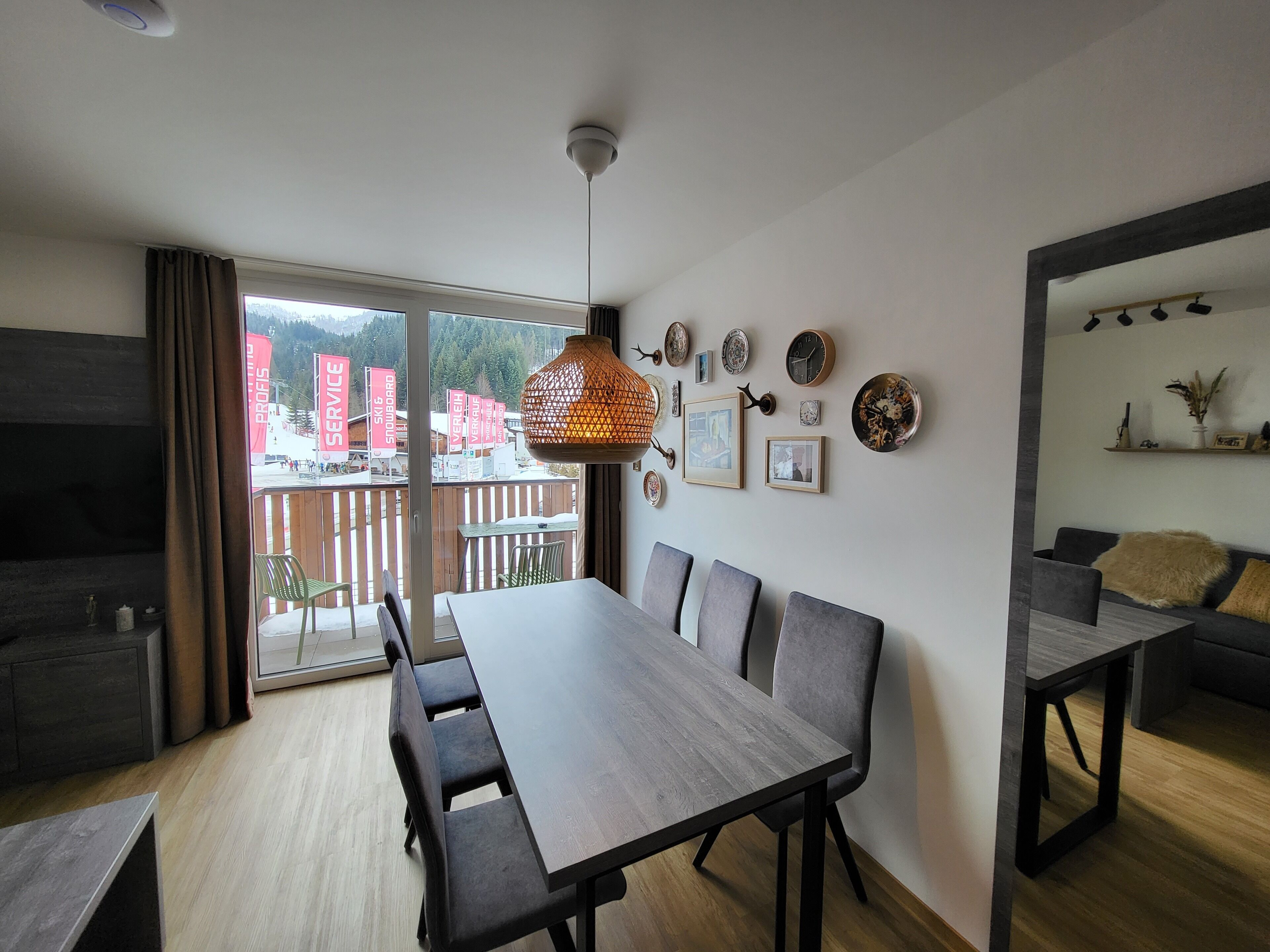 A modern dining area with a long table and chairs, a hanging light fixture, and a view of a snowy outdoor area through large windows.
