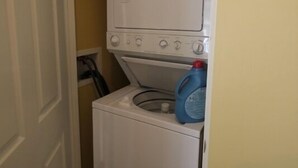 Un lavadero pequeño con lavadora y secadora de carga frontal, una botella de detergente a la vista y una puerta que da a otra habitación.