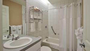 Baño con lavamanos, inodoro y bañera. Hay repisas sobre el lavamanos y una cortina de ducha.