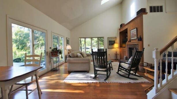 A spacious living room with a wooden floor, a sofa, a rocking chair, and a dining table. Large windows allow natural light to fill the space.