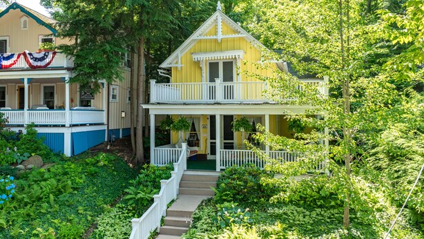 Une maison jaune à deux étages, dotée d’un porche et d’un balcon blancs, et entourée d’une végétation luxuriante.