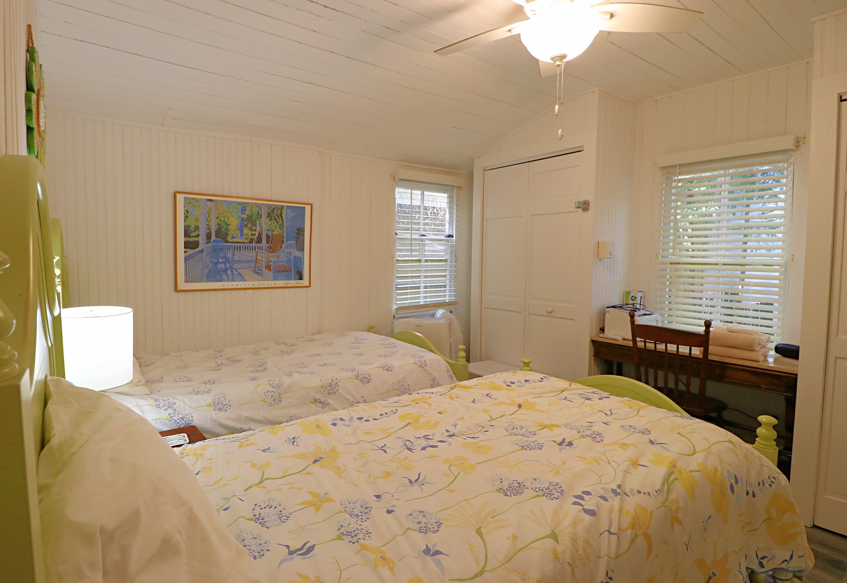 A bedroom with two beds, a ceiling fan, a painting on the wall, and a window with blinds.