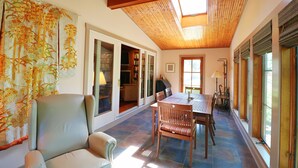 Una habitación iluminada por el sol, con techo de madera, una mesa de comedor con sillas, un sofá y una estantería.