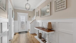 A hallway with a wooden floor, white walls, a white door with a glass panel, a staircase, and a console table with decorative items. - Discover Oak Island Nest Egg: a spacious 5-bedroom retreat with ocean views and private pool. (Oak Island)