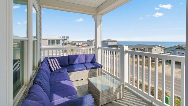 A covered balcony with a blue sofa and a glass-top table, overlooking a coastal road and buildings. - Discover Oak Island Nest Egg: a spacious 5-bedroom retreat with ocean views and private pool. (Oak Island)