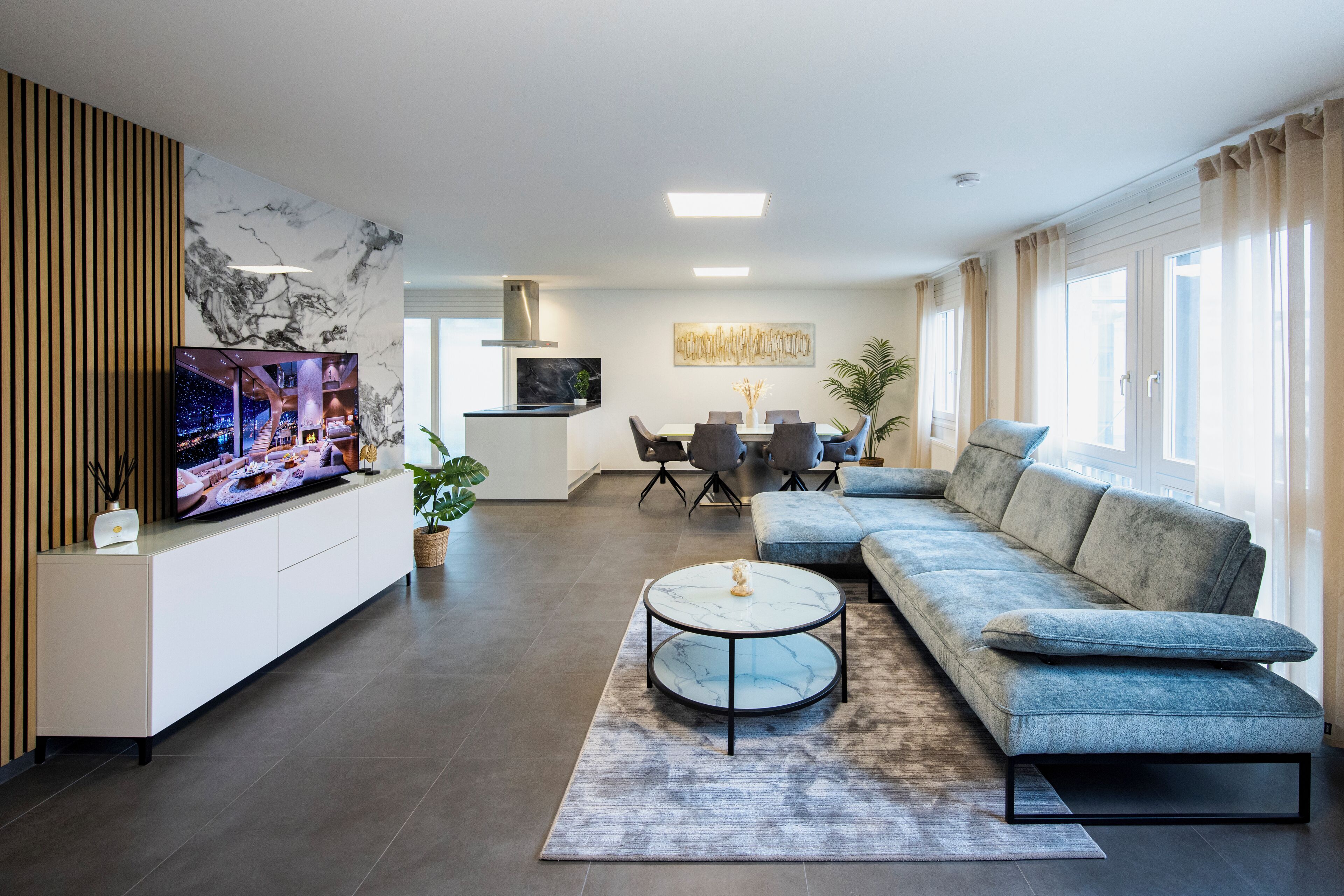 Ein modernes Wohnzimmer mit einem Ecksofa, einem runden Couchtisch, einem Flachbildfernseher und einem Essbereich mit Tisch und Stühlen.