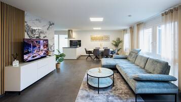 Ein modernes Wohnzimmer mit einem Ecksofa, einem runden Couchtisch, einem Flachbildfernseher und einem Essbereich mit Tisch und Stühlen.