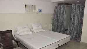 Cama doble con ropa de cama blanca, una silla de madera, una mesita pequeña, un equipo de aire acondicionado y una cortina de ducha con estampado floral.