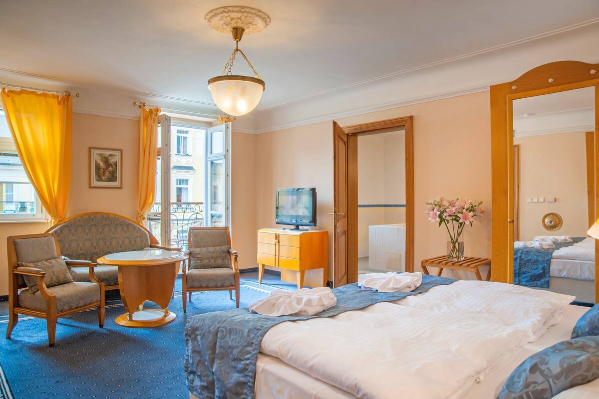 A hotel room with a bed, two armchairs, a small table, a television, and a mirror.