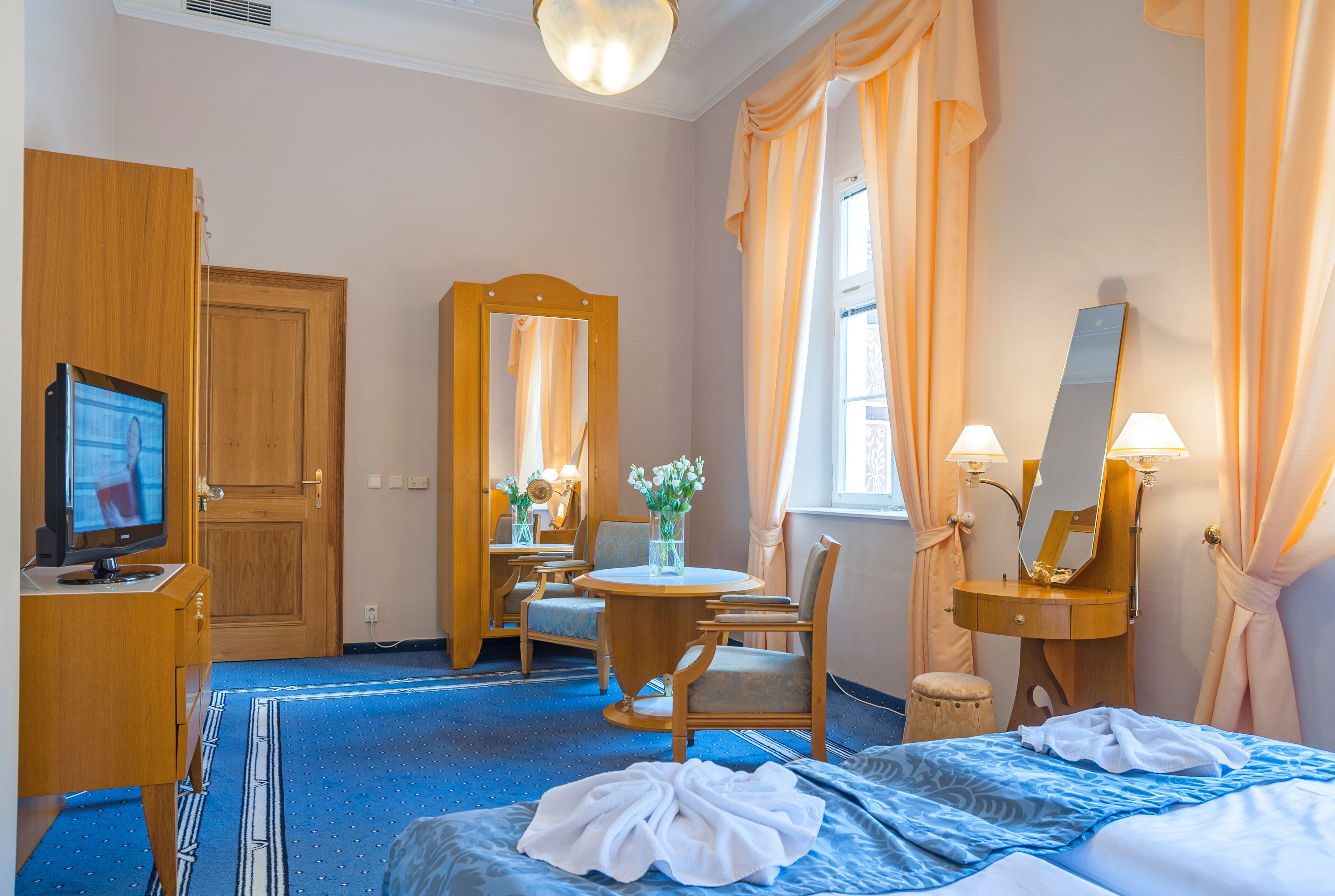 A hotel room with a bed, a desk with a computer, a chair, a round table, a mirror, and a window with curtains.