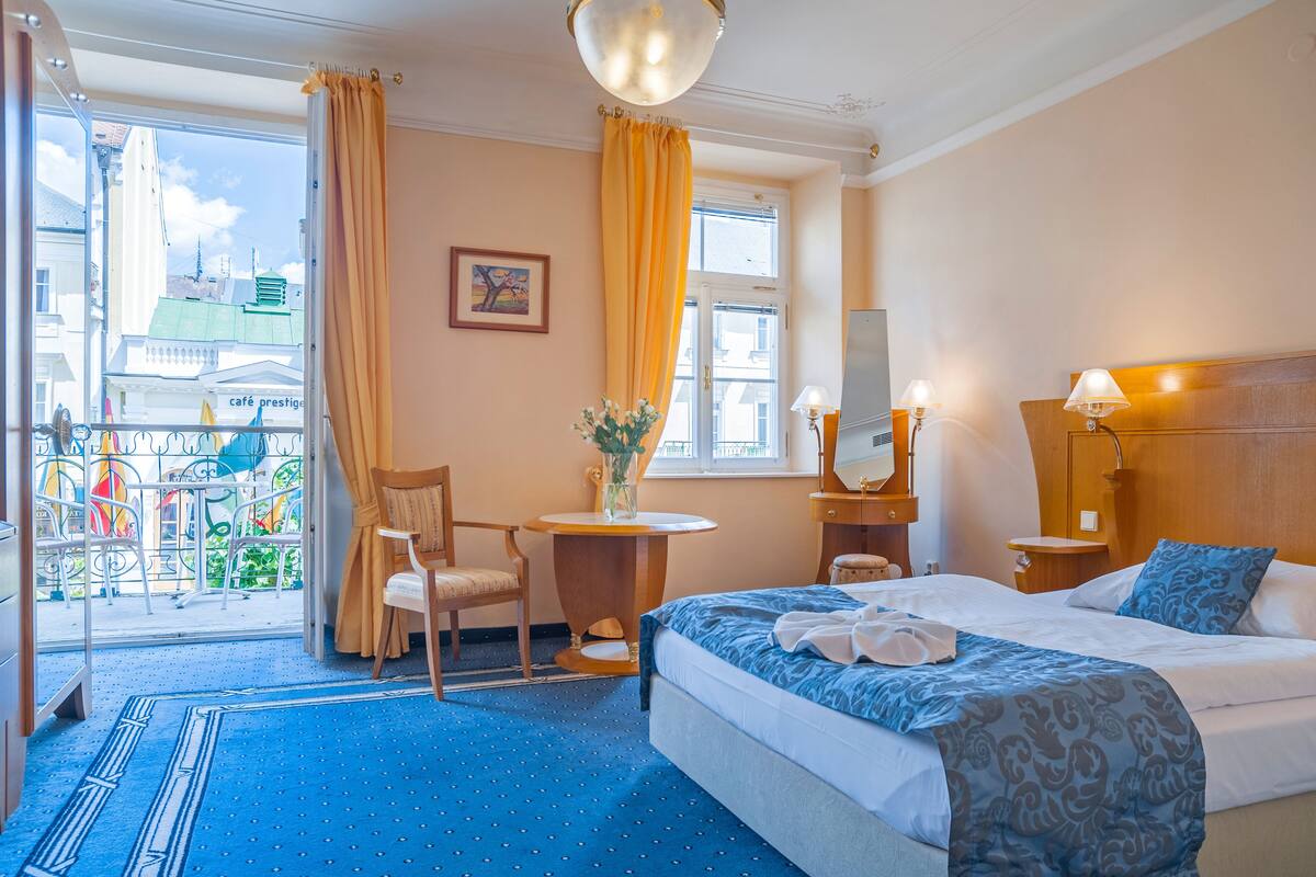 A hotel room with a bed, a desk, a chair, a small table with a vase, and a view of the city through a window.