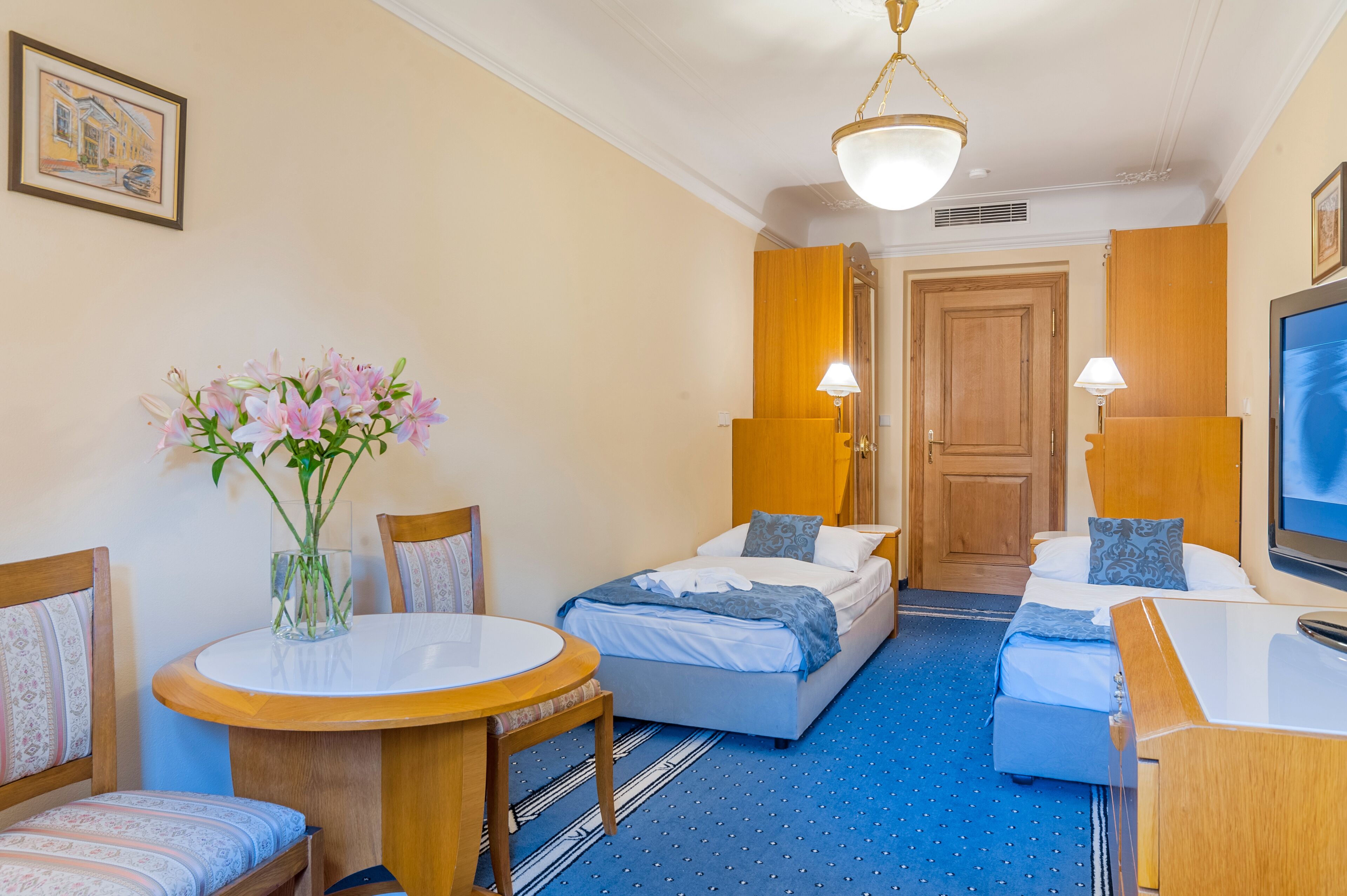 A hotel room with a bed, a desk, a chair, a table, and a vase of flowers.