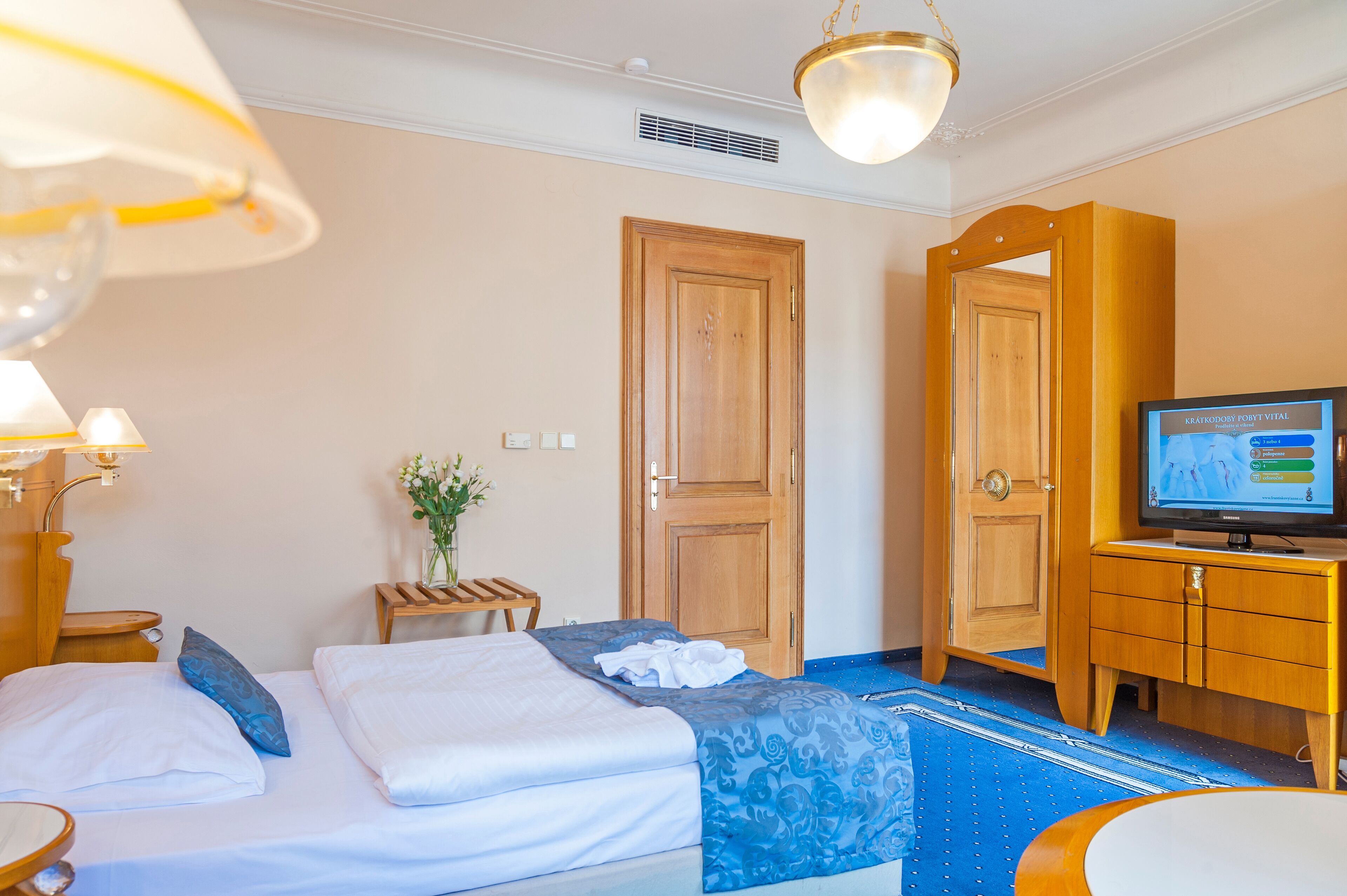 Una habitación de hotel con cama, televisión, armario de madera y una mesita con un jarrón de flores.