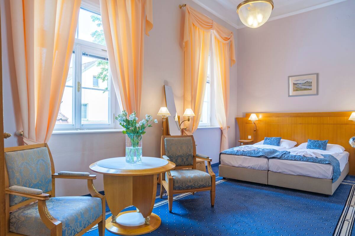 A hotel room with a bed, two chairs, a round table with a vase, and a painting on the wall.