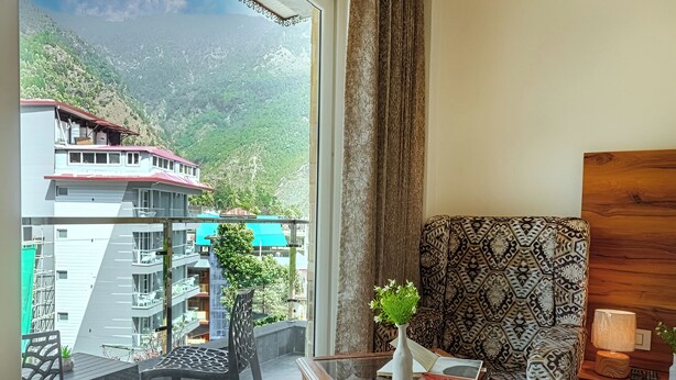 A hotel room with a large window offering a view of a mountainous landscape, a patterned armchair, a small wooden table, and a nightstand with a lamp. - Hotel Classic Hills (Dharamshala)