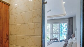 A bathroom with a marble wall, a wooden cabinet, and a large mirror reflecting a bedroom with a bed and a window. - Hotel Classic Hills (Dharamshala)