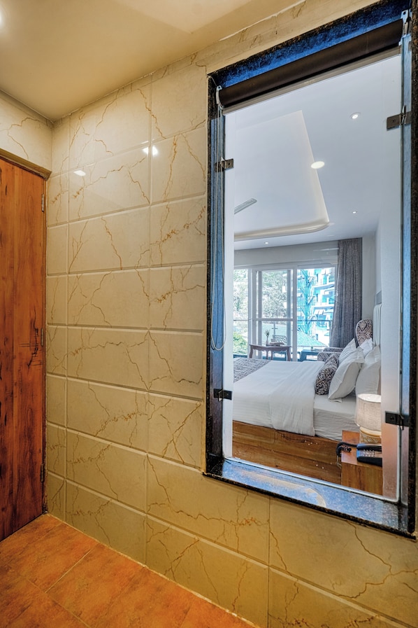 Un baño con pared de mármol, un mueble de madera y un espejo grande que refleja un dormitorio con cama y ventana.
