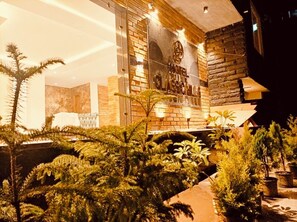 A hotel entrance with a stone facade, a large illuminated sign, and potted plants in the foreground. - Hotel Classic Hills (Dharamshala)