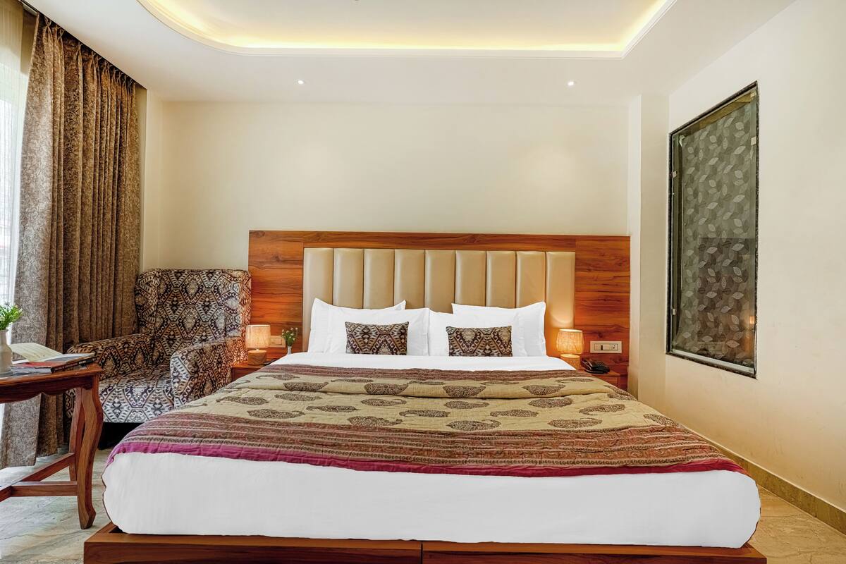 A hotel room with a large bed, a wooden headboard, patterned pillows, a chair, bedside lamps, and a large abstract painting on the wall.