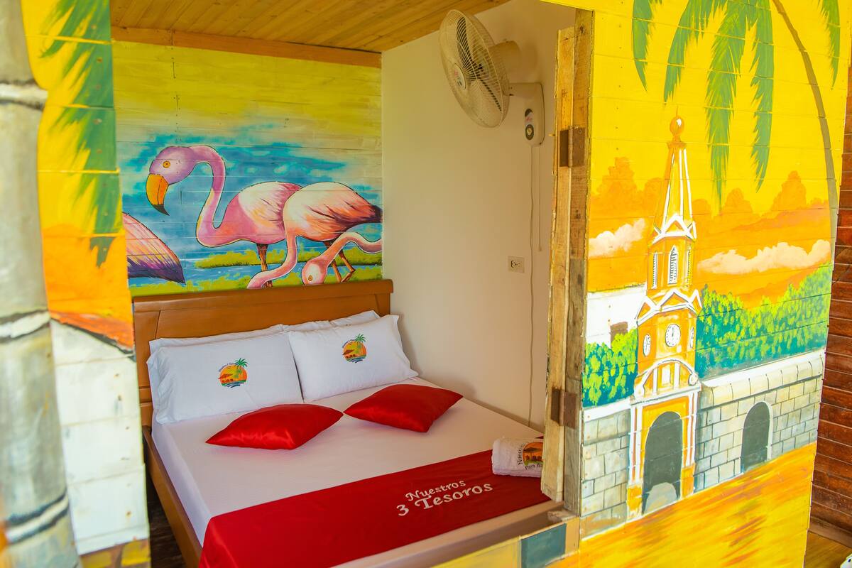 A hotel room with a bed, a mural of a flamingo and a church, and a fan on the wall.