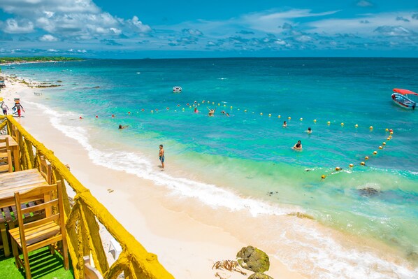 A beach with a wooden deck and chairs, clear turquoise water, and a few people swimming.