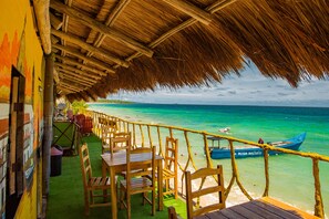 A beachside restaurant with wooden tables and chairs, a thatched roof, and a view of the ocean and a boat.