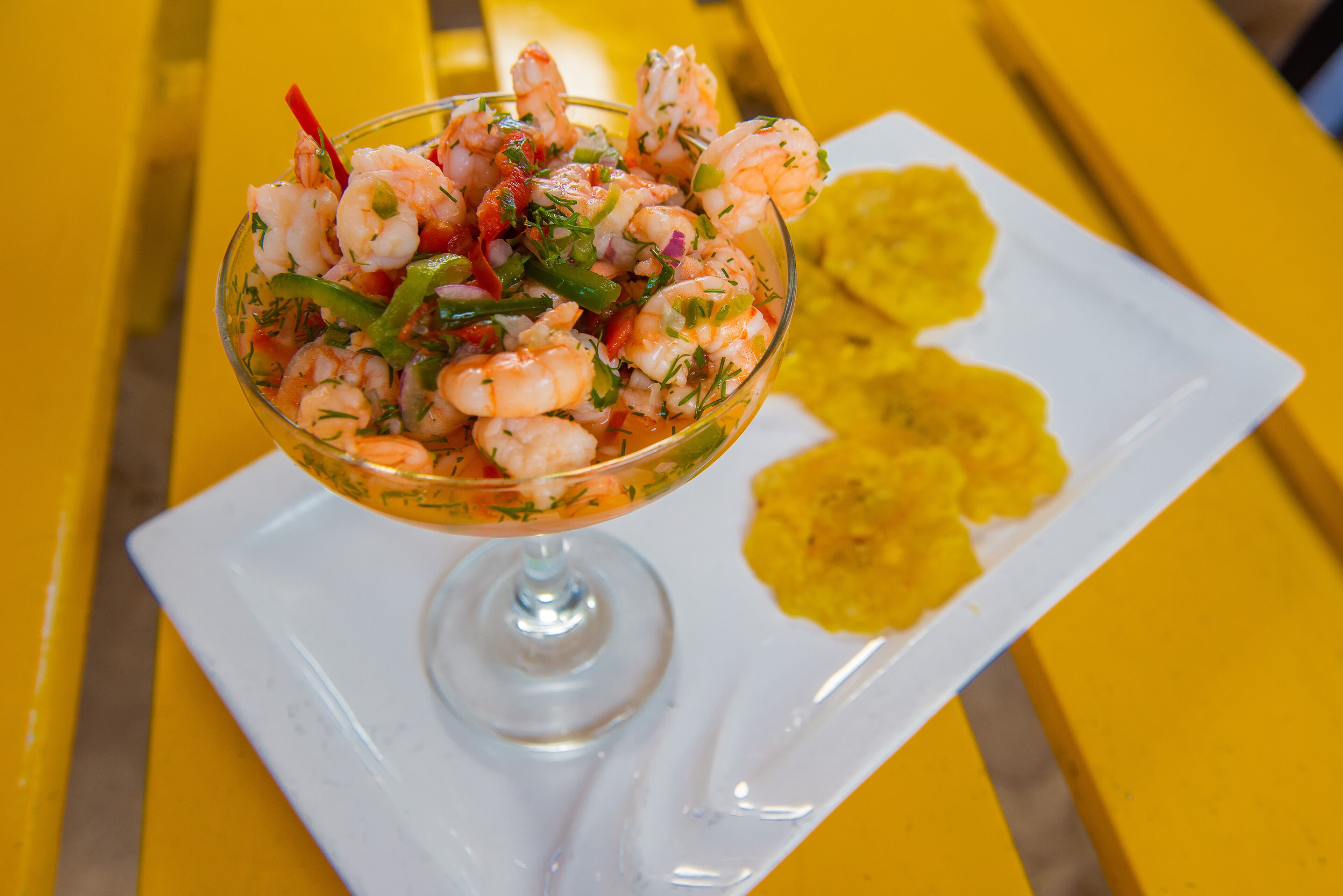 A shrimp cocktail in a glass bowl on a white plate, accompanied by yellow plantain chips, set on a yellow table.