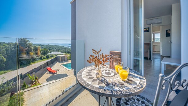 A balcony with a view of a swimming pool and greenery, a table with a vase and glasses of orange juice, and a modern interior with a white wall and a door. - Villa Rozeta with sea view (Šegotići)