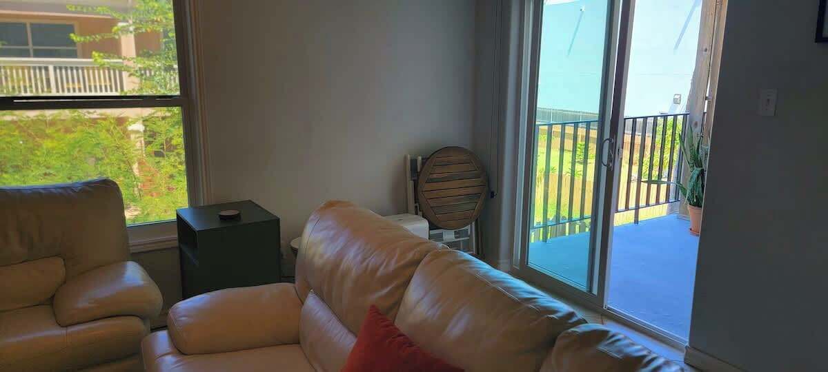 A living room with a beige leather sofa, a small side table, and a wooden chair. A large window offers a view of a balcony with a railing and some greenery.